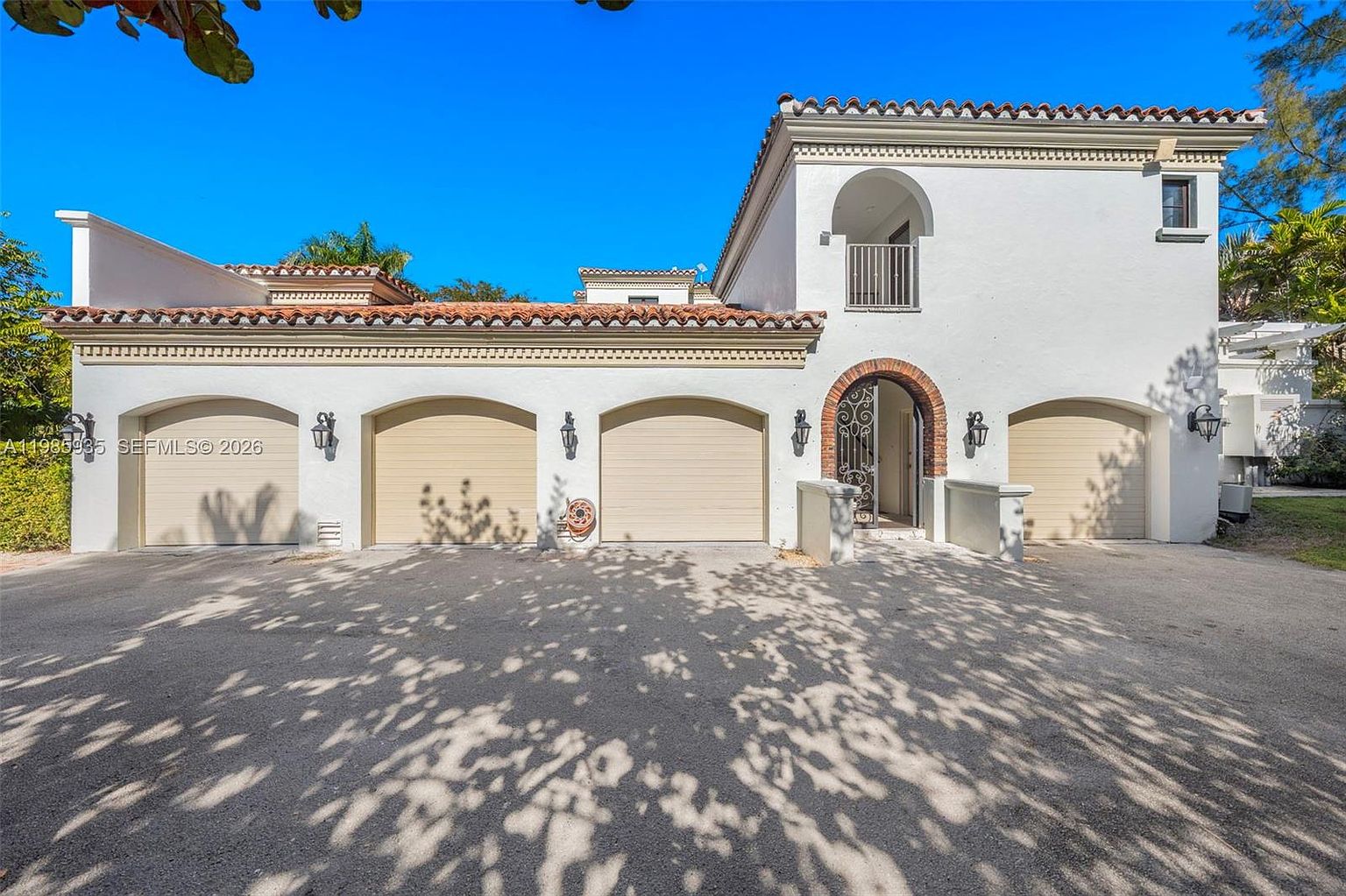 This Mediterranean-style home features a prominent white stucco facade with a multi-car garage and a distinctive arched entryway. The exterior is accented by a terracotta-tiled roof, decorative ironwork on the front door, and a small second-story balcony. The perspective is a wide-angle shot from the driveway, capturing the full width of the home under a clear blue sky.