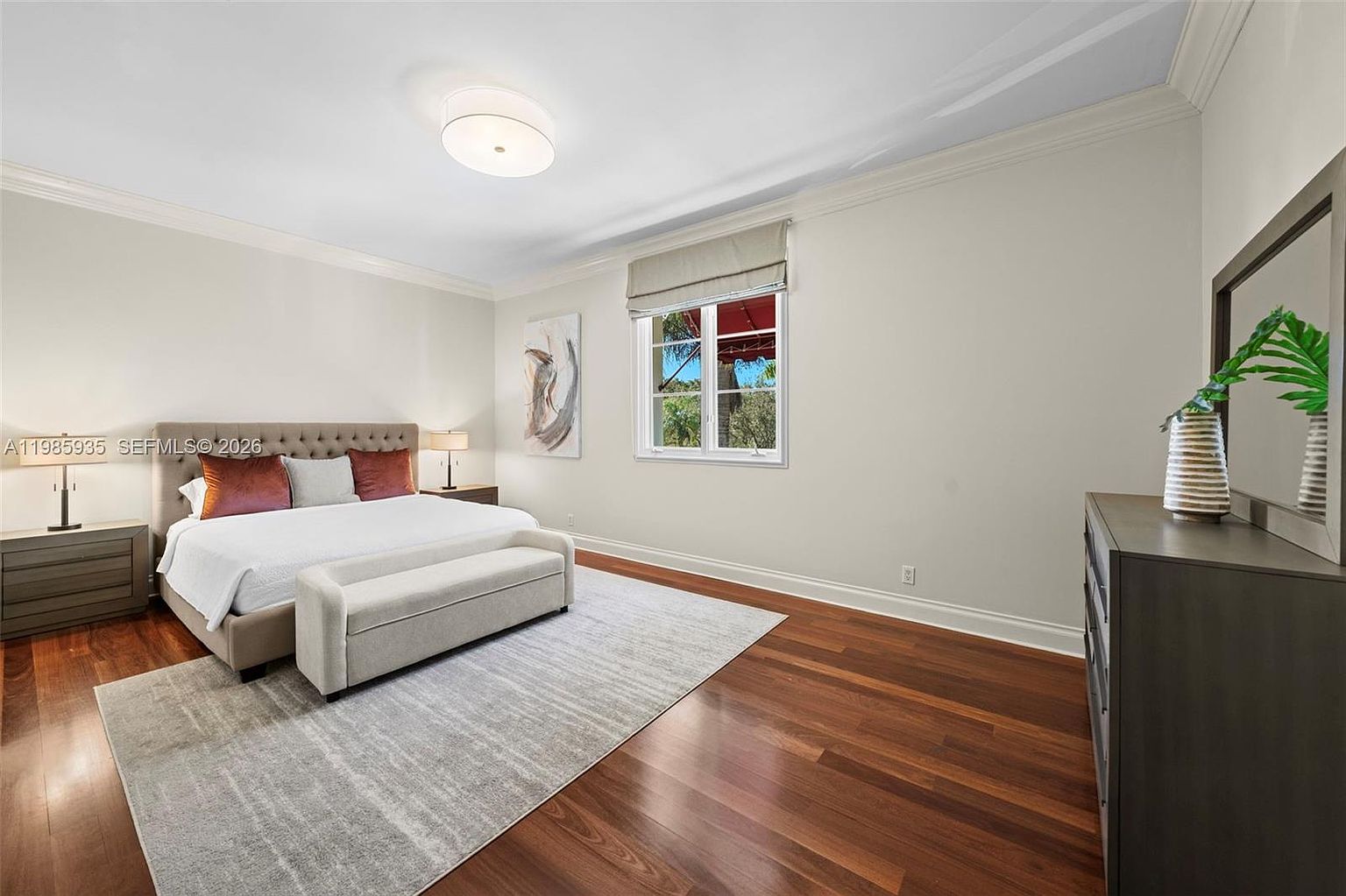 This spacious primary bedroom features rich, dark wood flooring and neutral-toned walls, creating a serene and sophisticated atmosphere. A large, tufted headboard anchors the bed, which is dressed in crisp white linens and accented with warm-toned pillows, while a matching bench sits at the foot. The room is illuminated by a central flush-mount light fixture and includes a large dresser with a mirror, all framed by a window that allows natural light to brighten the space.