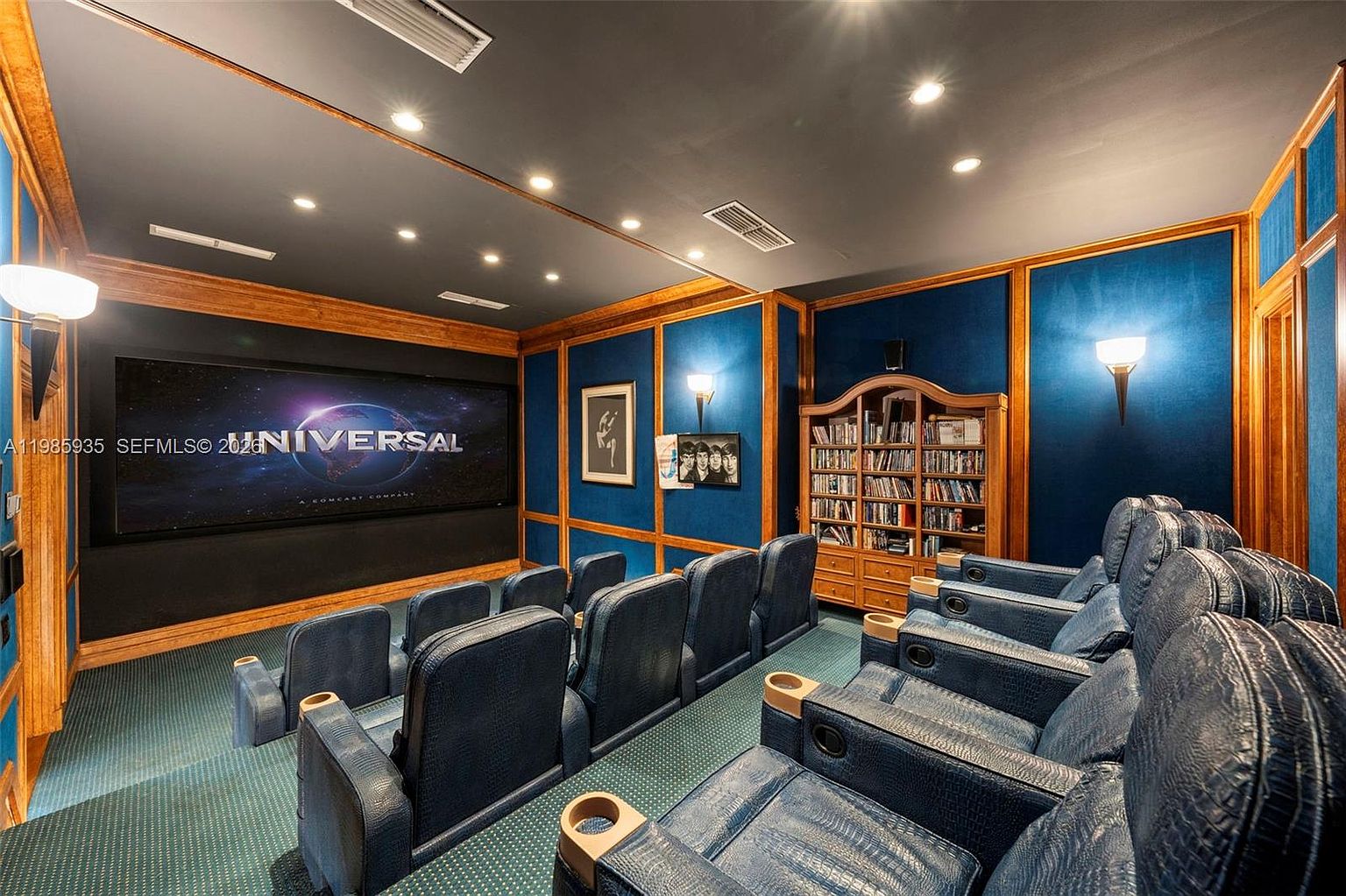 This dedicated home theater features tiered rows of blue leather seating arranged for optimal viewing of a large projection screen. The room is accented with wood-paneled walls, blue fabric wall treatments, and recessed ceiling lighting, creating a classic cinematic atmosphere. A built-in wooden bookcase provides storage for media, and the space is carpeted throughout to enhance the viewing experience.