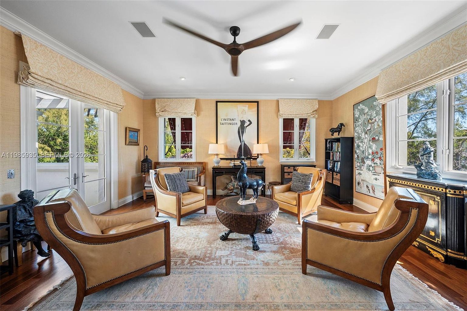 This elegant living room features a symmetrical layout with four leather armchairs arranged around a central circular wooden coffee table. The space is adorned with warm-toned wallpaper, a large patterned area rug, and a prominent ceiling fan, creating a sophisticated and inviting atmosphere. Large windows with fabric shades allow natural light to fill the room, highlighting the curated decor and artwork.