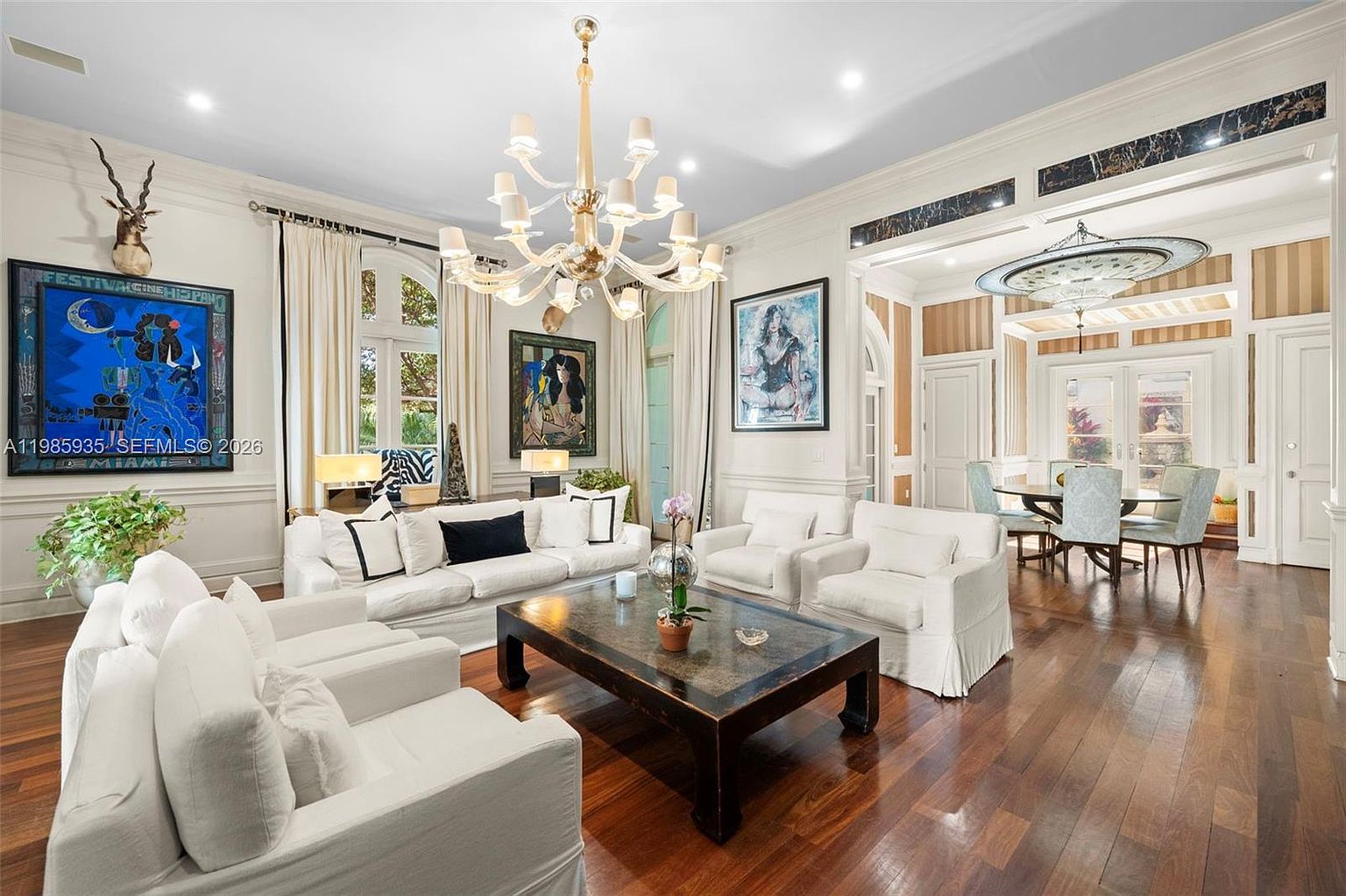 This elegant living room features rich, dark wood flooring and a sophisticated neutral color palette with white upholstered seating. A grand, multi-tiered chandelier serves as the focal point, complemented by vibrant, large-scale artwork on the walls and tall windows draped in floor-length curtains. The open-concept layout flows seamlessly into an adjacent dining area, creating a spacious and refined atmosphere suitable for formal entertaining.