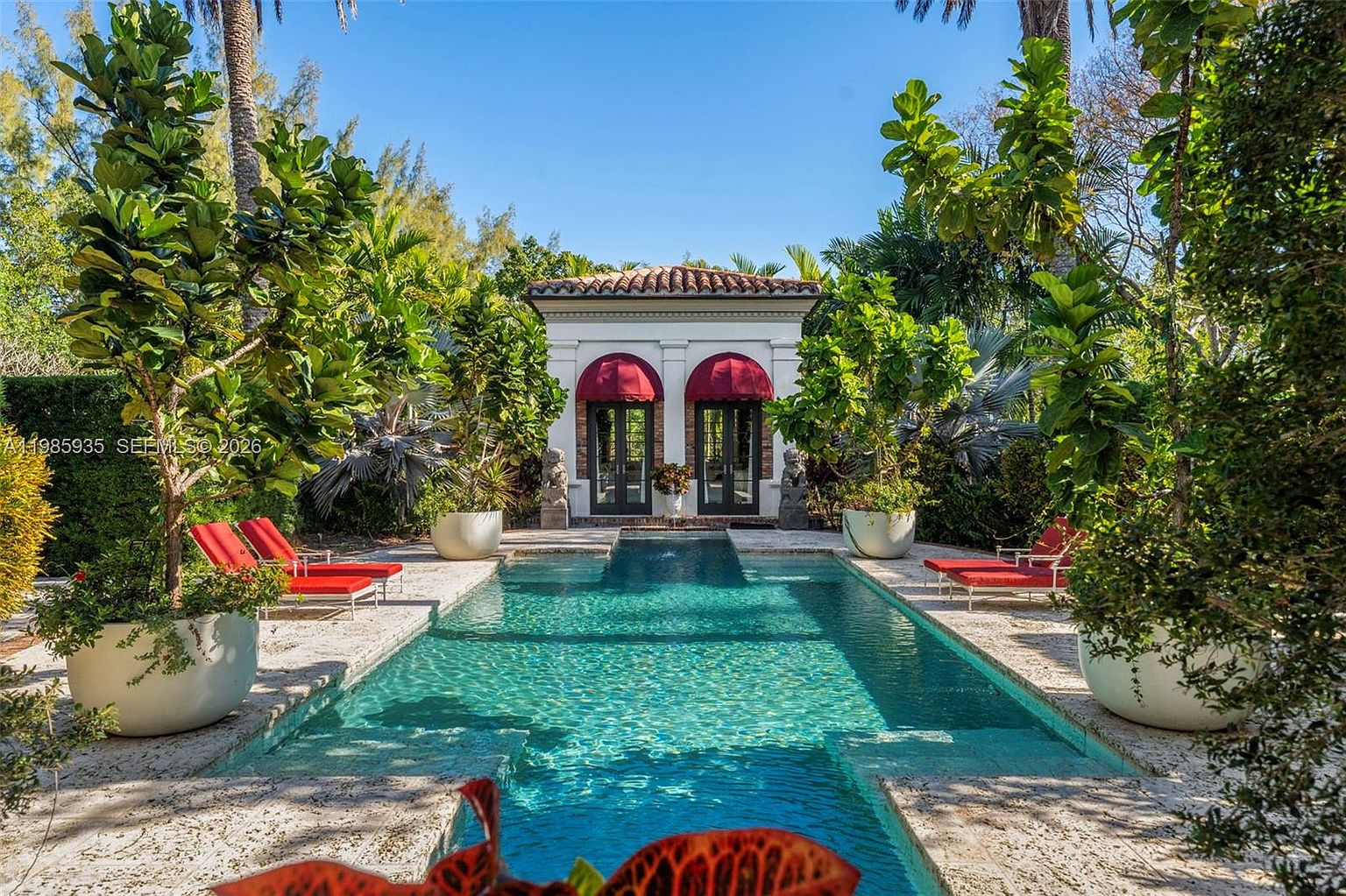 This elegant outdoor space features a rectangular swimming pool centered in a lush, tropical garden setting. A classic pavilion with red fabric awnings and arched glass doors serves as a focal point at the far end of the pool, flanked by stone statues. The surrounding stone patio is furnished with vibrant red lounge chairs, creating a sophisticated and serene retreat.
