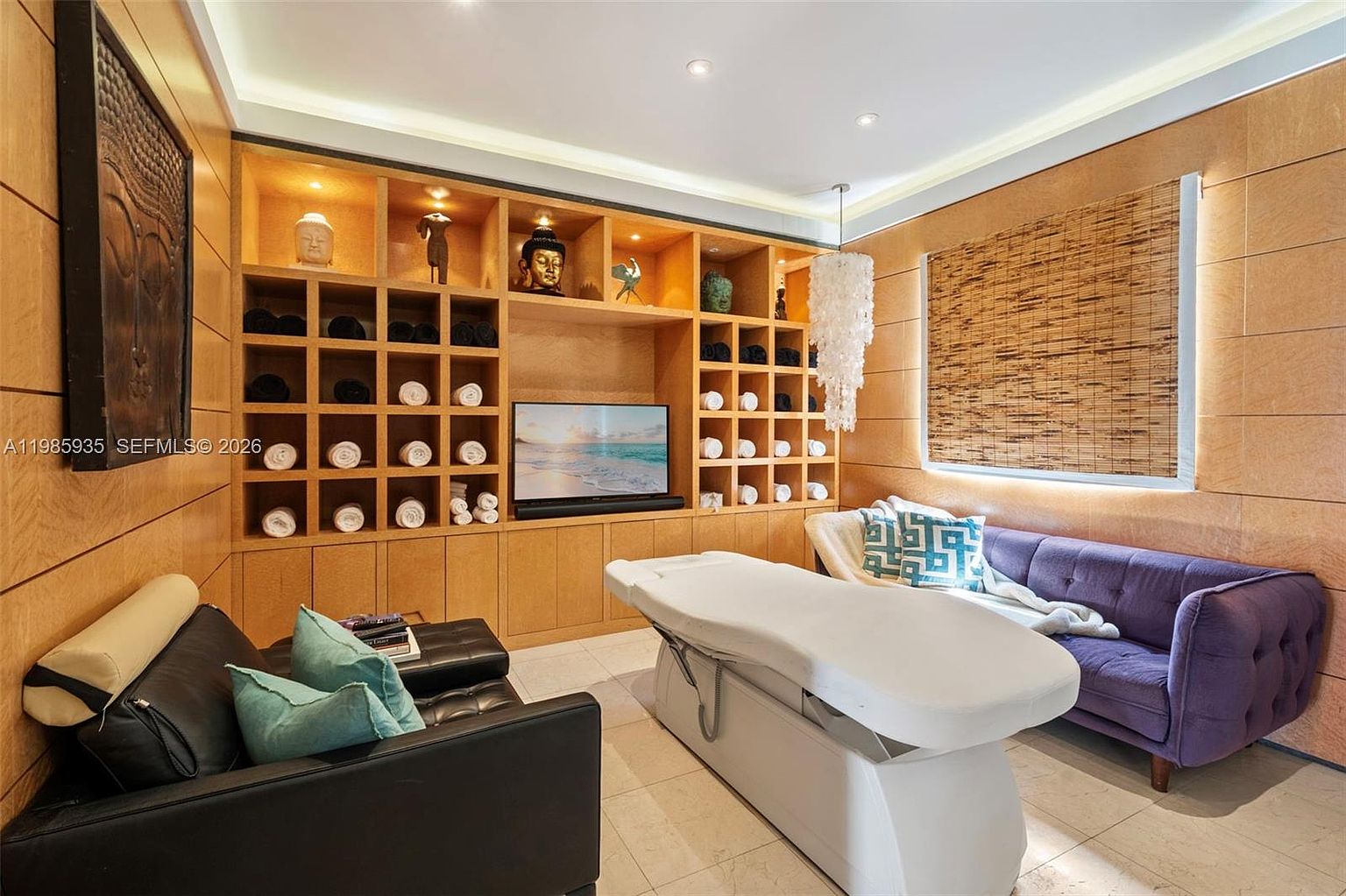 This dedicated spa or massage room features a professional treatment table as the central focus, flanked by a purple tufted sofa and a black leather lounge chair. A large, light wood built-in shelving unit covers the back wall, displaying rolled towels and decorative Buddha-themed sculptures alongside a flat-screen television. The room is accented by warm ambient lighting, a unique shell-style chandelier, and a bamboo-style window shade, creating a serene and tranquil atmosphere.