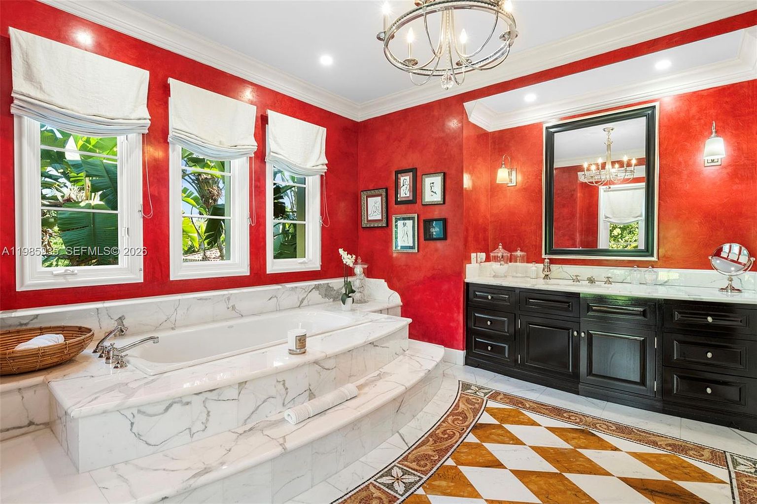 This primary bathroom features bold red walls contrasted by a large, white stone-clad soaking tub set on a raised platform. A dark wood vanity with a light-colored stone countertop and a large mirror occupies the right side of the room, while a patterned tile floor adds visual interest. Three large windows with white shades allow natural light to fill the space, which is anchored by a traditional chandelier.