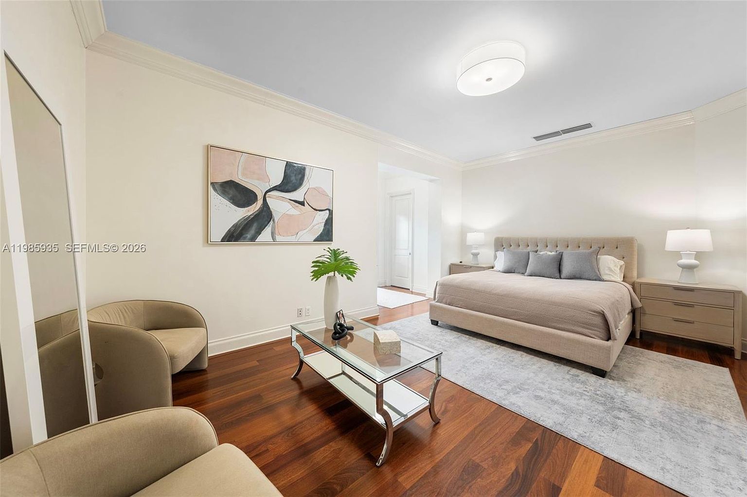 This spacious primary bedroom features rich, dark wood flooring and a neutral color palette that creates a serene atmosphere. The room is anchored by a large, upholstered bed with a tufted headboard, flanked by matching nightstands and lamps, while a glass-topped coffee table and two armchairs provide a comfortable seating area. A large abstract painting hangs above the seating area, and the room is illuminated by a modern, circular flush-mount ceiling light.