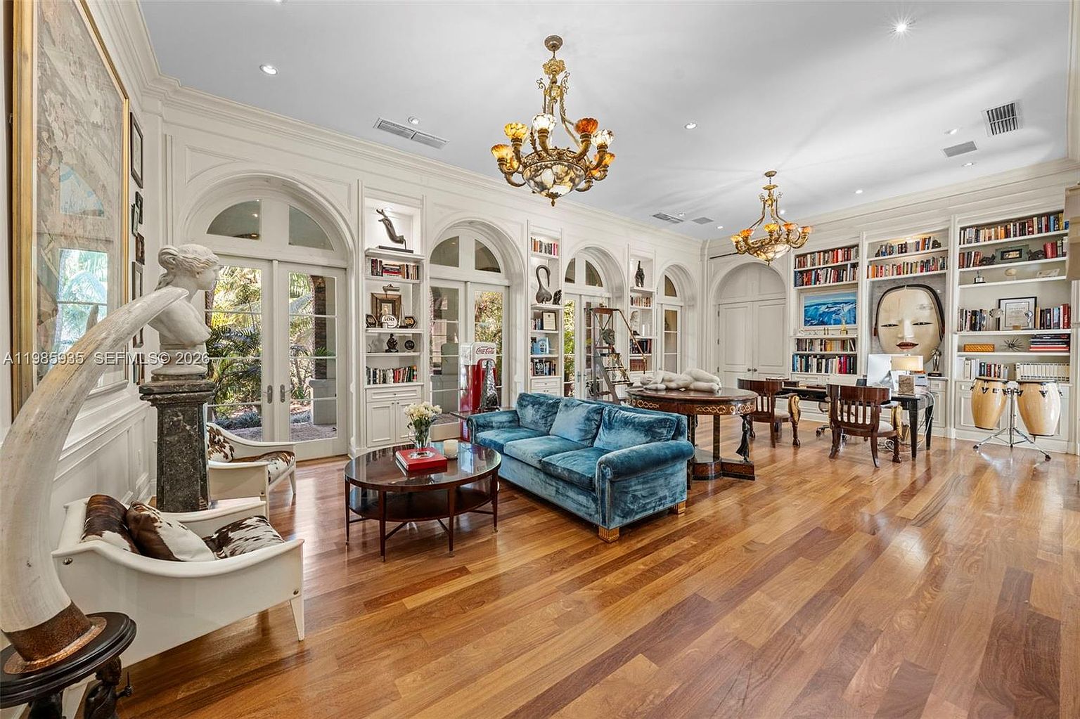 This expansive, elegant living room features rich wood flooring and tall, arched glass doors that open to the outdoors, flooding the space with natural light. The room is furnished with a vibrant blue velvet sofa, a circular coffee table, and two patterned armchairs, all accented by ornate chandeliers and built-in shelving units filled with books and decor. A large, striking mask sculpture serves as a focal point near the office area, creating a sophisticated and eclectic atmosphere.