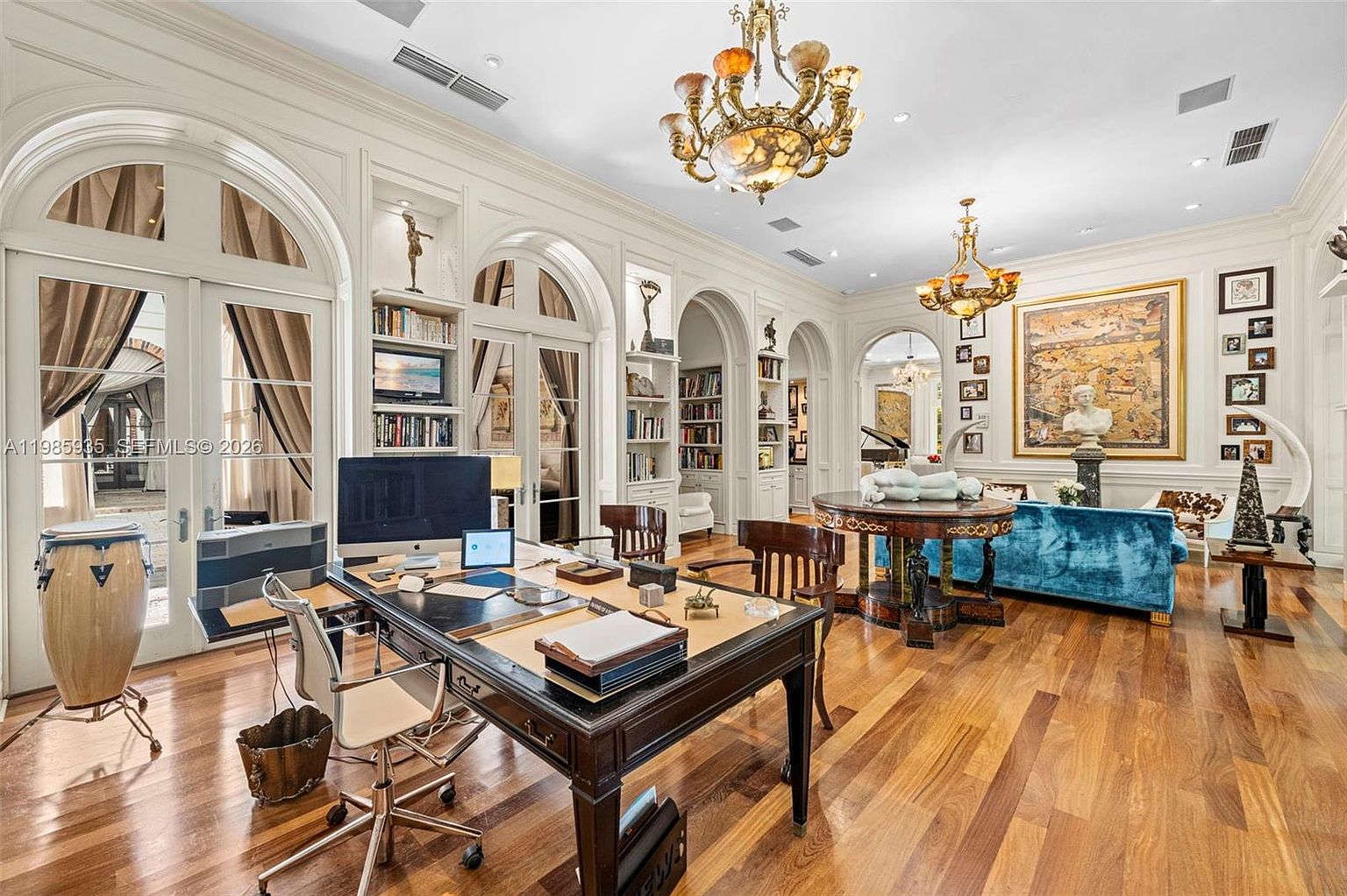 This expansive, classically inspired home office features rich wood flooring and high ceilings adorned with ornate chandeliers. The room is lined with built-in shelving and tall arched glass doors that provide natural light, while a large dark wood desk serves as the central workspace. A vibrant blue sofa and a circular pedestal table create a sophisticated seating area, complemented by curated artwork and decorative sculptures throughout the space.