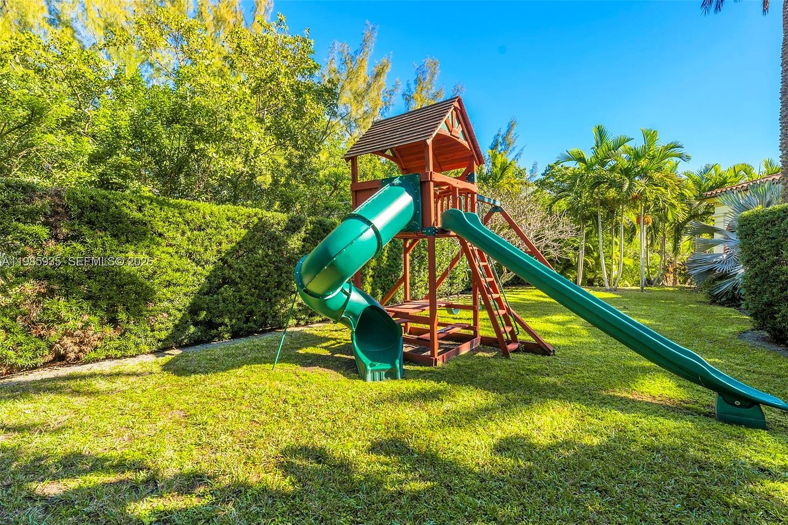 This image captures a lush, green backyard featuring a wooden playset with a slide and a climbing structure. The yard is enclosed by dense, well-manicured hedges and mature palm trees, creating a private and serene outdoor space. The bright, sunny lighting highlights the vibrant lawn and the inviting nature of this family-friendly area.