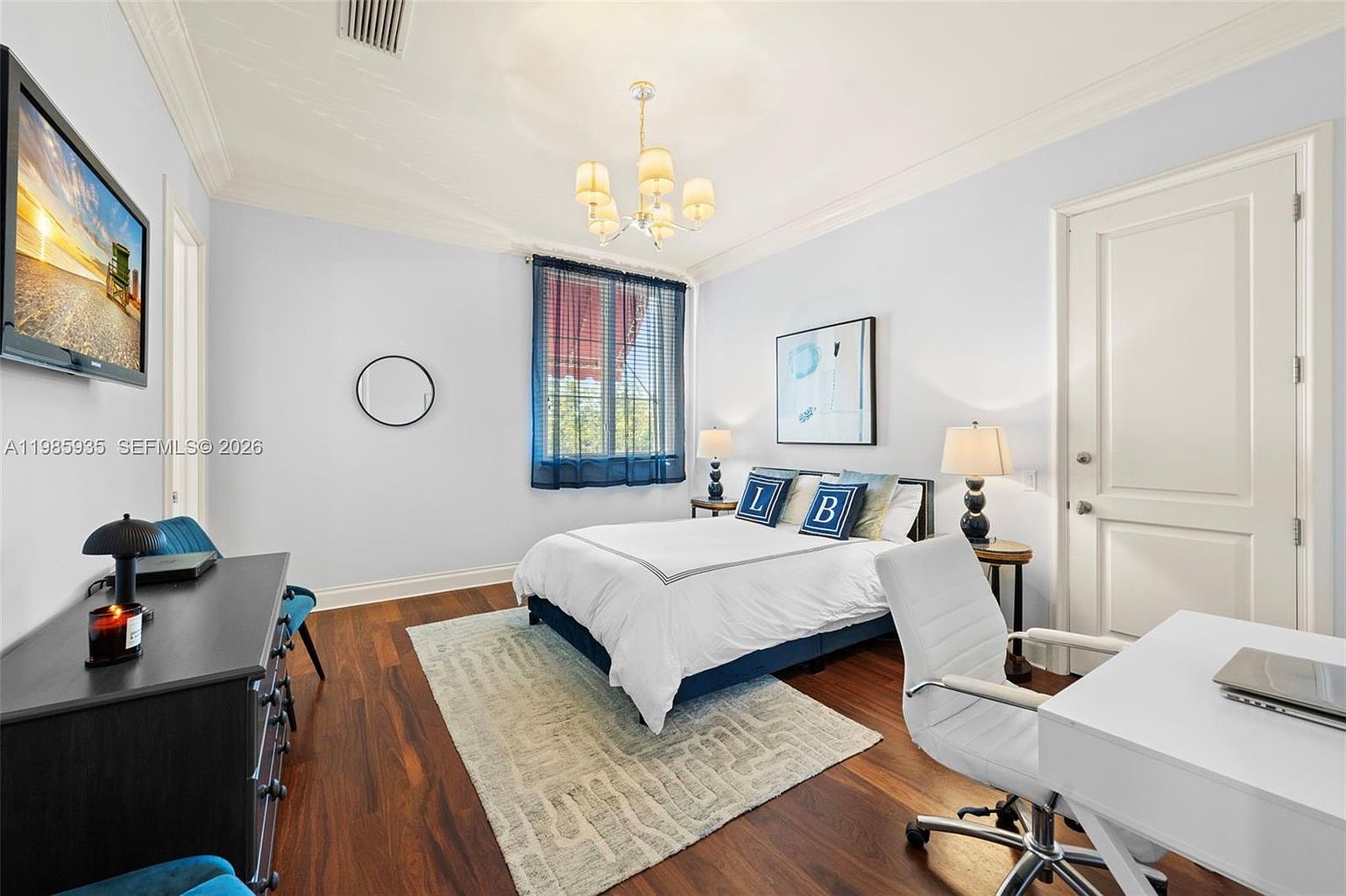 This well-lit bedroom features dark wood flooring and light-colored walls, creating a balanced and inviting atmosphere. The room is furnished with a central bed adorned with decorative pillows, flanked by two matching nightstands with lamps, and includes a dedicated workspace with a white desk and chair. A large television is mounted on the wall, and a window with blue curtains allows natural light to brighten the space.