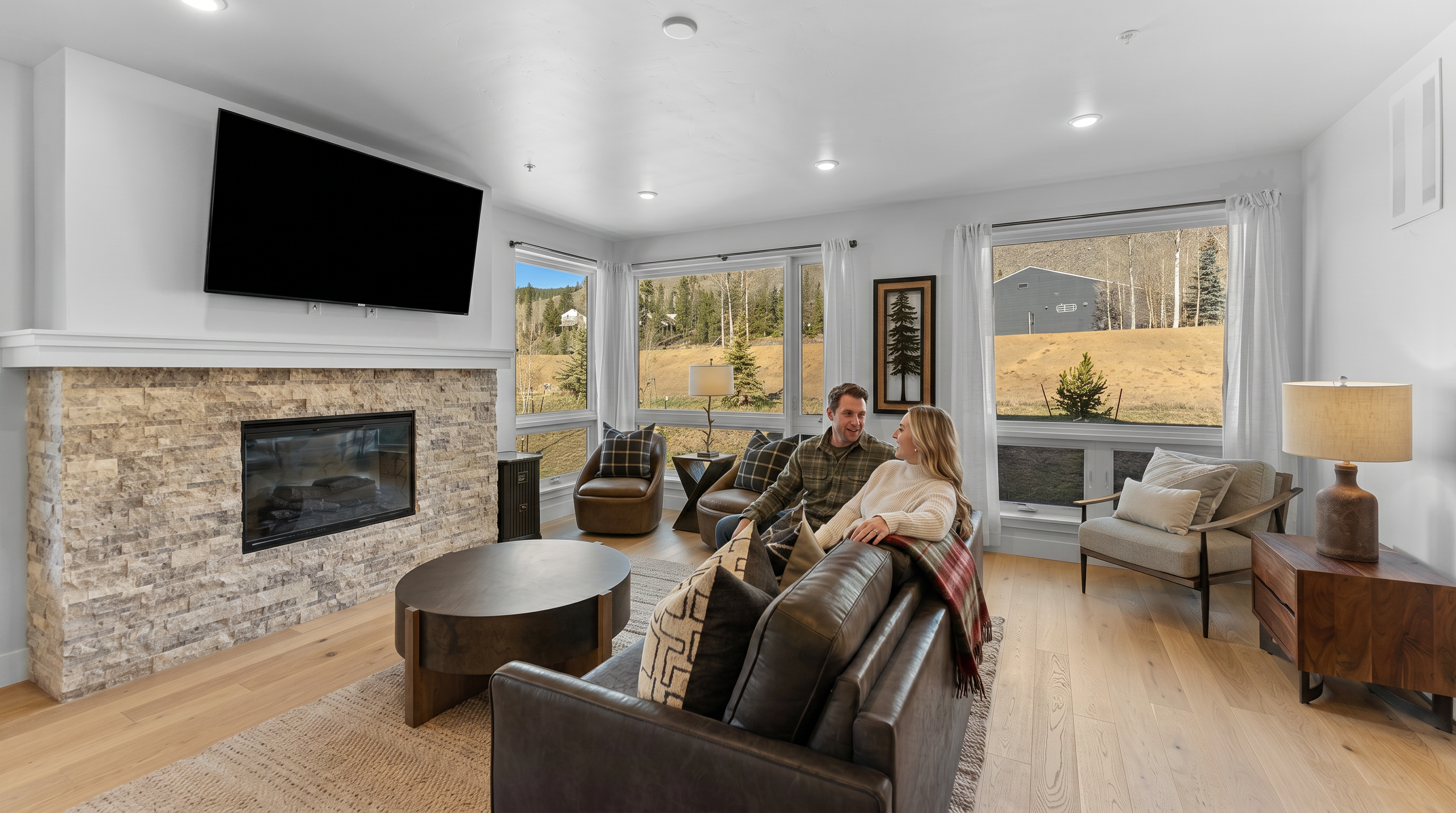 This inviting living room features a prominent stone fireplace with a mounted television, creating a cozy focal point. The space is furnished with a dark leather sofa, a round coffee table, and additional armchairs, all arranged on a textured area rug over light hardwood floors. Large windows provide ample natural light and scenic views of the surrounding landscape, contributing to a warm, mountain-modern aesthetic.