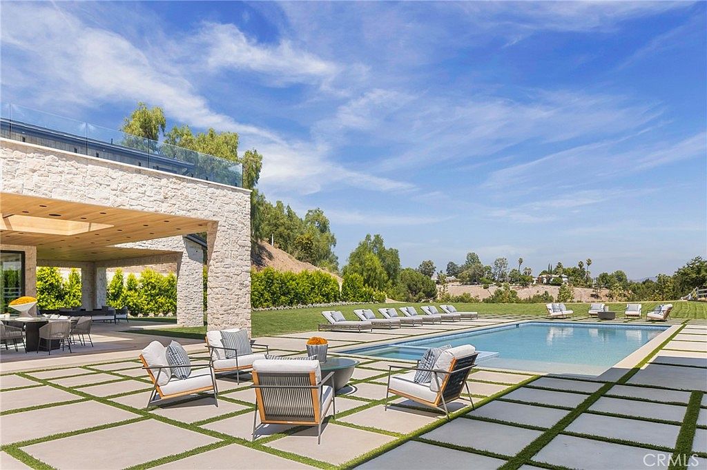 This image showcases a luxurious backyard with a large rectangular pool as its centerpiece. The pool is surrounded by a paved patio area with modern outdoor furniture, including lounge chairs and seating arrangements. The property features well-maintained landscaping, including lush greenery and mature trees, creating a private and serene outdoor living space.