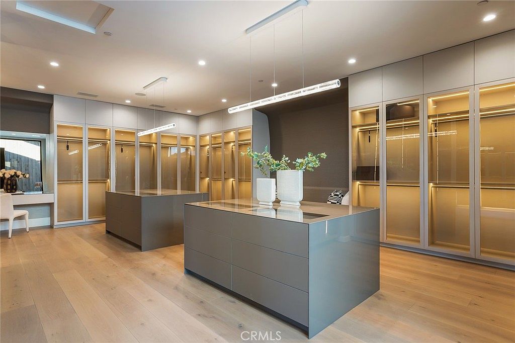 This is a spacious walk-in closet featuring custom cabinetry with glass doors and integrated lighting. A large center island with drawers provides additional storage and countertop space, while light wood flooring adds warmth to the room. The closet is well-organized and designed for luxury living.