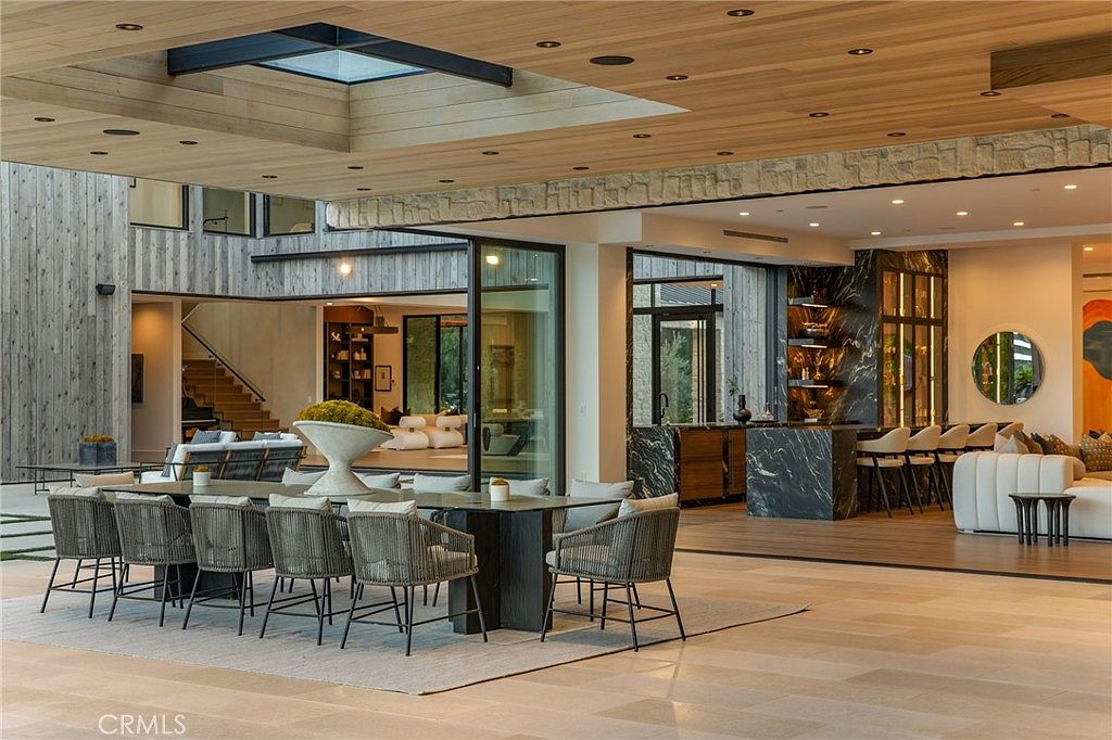 This interior shot showcases a luxurious dining area with a large table surrounded by woven chairs, set on a neutral rug. The open-concept design seamlessly connects to a bar area with marble accents and a comfortable seating area, all under a striking wood-paneled ceiling with a skylight. The space exudes modern elegance and sophistication.