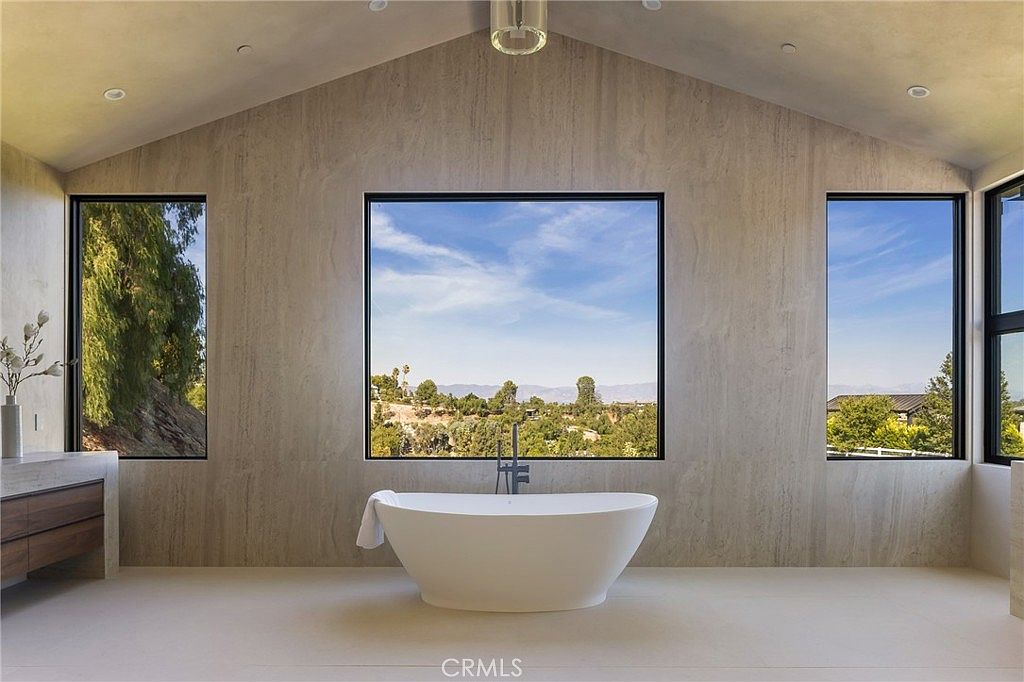 This is a luxurious primary bathroom featuring a freestanding white bathtub positioned in front of a large window with a scenic view. The walls are covered in a light-colored, textured material, and the room has a minimalist design with clean lines and a vaulted ceiling. The overall impression is one of serenity and high-end comfort.