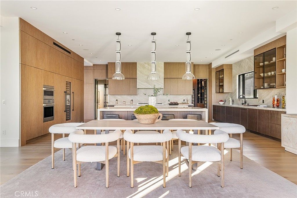 This is a bright and spacious kitchen and dining area featuring modern design elements. The kitchen boasts sleek wooden cabinetry, stainless steel appliances, and a large island, while the dining area includes a long table surrounded by stylish white chairs on a light-colored rug. Three pendant lights hang above the dining table, adding to the contemporary aesthetic.