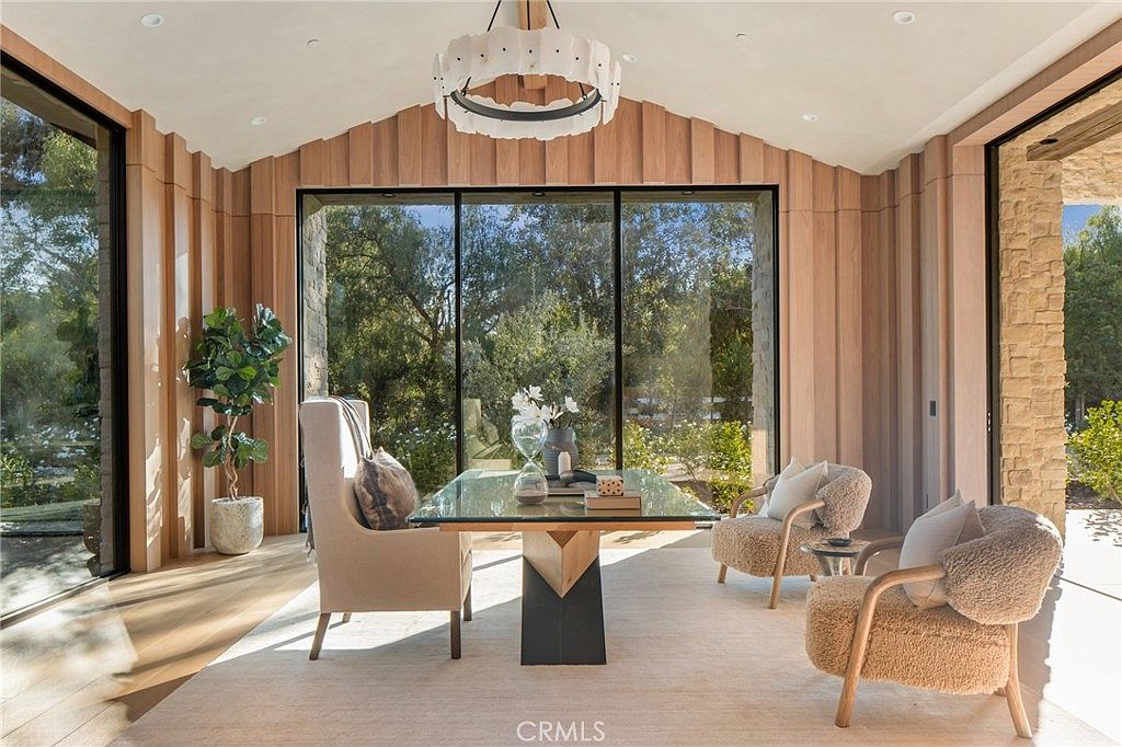 This is an interior shot of a modern office or study featuring a glass-top desk with a unique geometric base, surrounded by two plush armchairs and a high-backed chair. The room is characterized by natural wood paneling, a large window offering a view of lush greenery, and a contemporary chandelier, creating a bright and inviting workspace.
