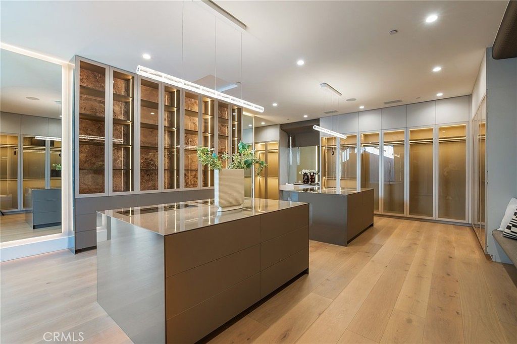 This is a spacious walk-in closet featuring custom shelving and storage solutions. The closet includes two central islands with drawers and reflective countertops, providing ample space for organization. The design incorporates modern lighting and a neutral color palette, creating a luxurious and functional space.