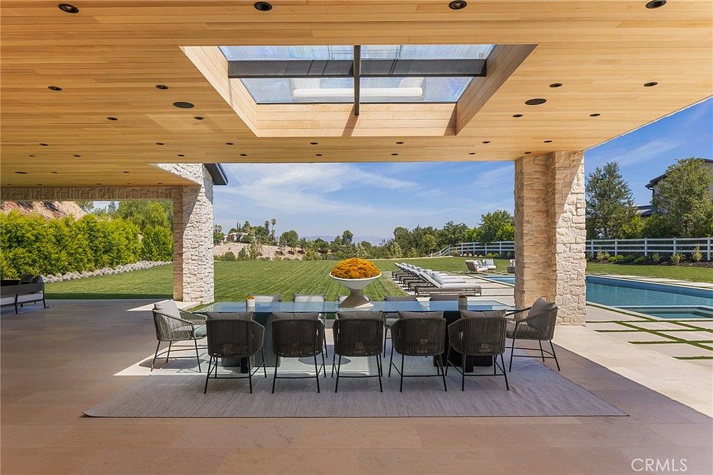 This image showcases a luxurious outdoor patio area, featuring a large dining table with seating for ten, set upon a neutral-toned rug. The patio is covered by a wooden ceiling with recessed lighting and a skylight, supported by stone pillars. Beyond the patio, a well-manicured lawn, a swimming pool, and lounge chairs create an inviting outdoor living space, all under a bright blue sky.