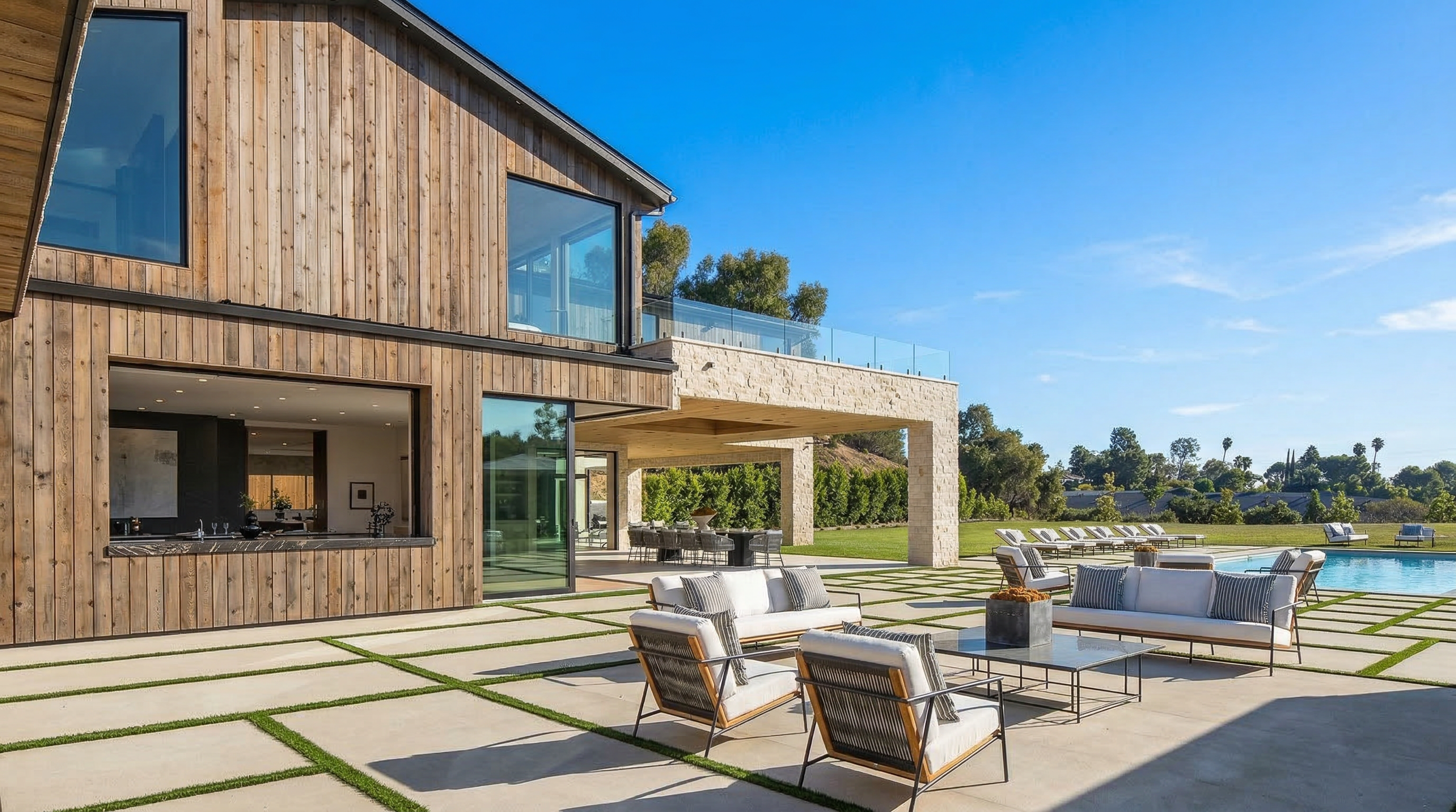 This modern luxury patio features an expansive concrete terrace with patterned artificial turf seams, providing an elegant outdoor living space. The architectural design combines natural wood siding with a stone-clad, covered patio area, which flows seamlessly into a resort-style backyard with a pool and lush landscaping. The setting is ideal for upscale outdoor entertaining, highlighting a sophisticated contemporary aesthetic.