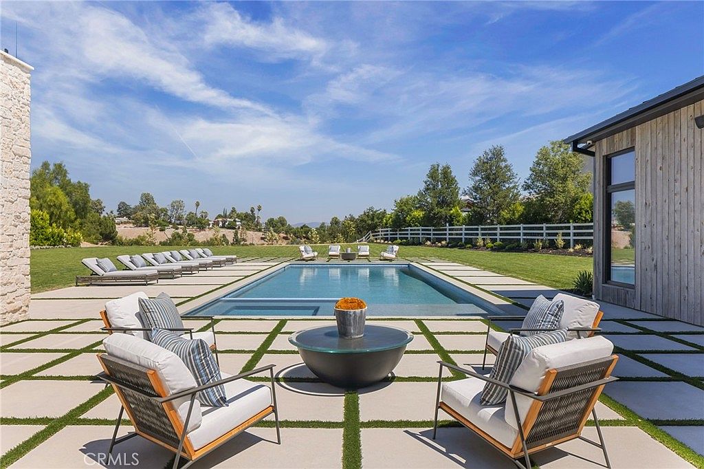 This image showcases a luxurious backyard pool area, featuring a rectangular pool surrounded by a modern grid of concrete pavers and artificial turf. Lounge chairs and a central fire pit with seating create an inviting space for relaxation and entertainment. The landscaping is well-maintained, and the overall aesthetic is clean, contemporary, and upscale.