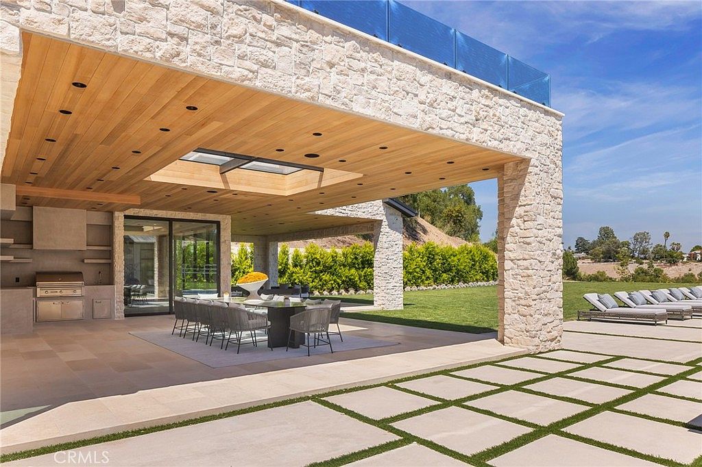 This image showcases a luxurious outdoor patio area, featuring a stone structure with a wooden ceiling and recessed lighting. A dining table with modern chairs sits on a large rug, while an outdoor kitchen area is visible in the background. The patio extends to a paved area with grass inserts, leading to lounge chairs, creating an inviting and sophisticated outdoor living space.