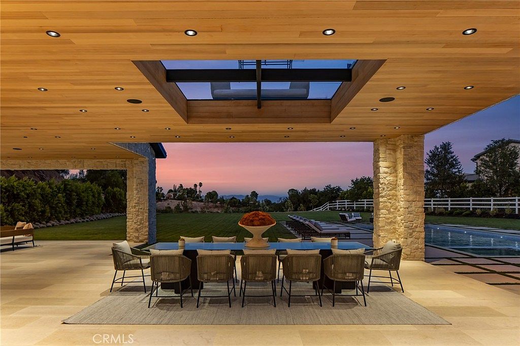 This image showcases a luxurious outdoor patio area, featuring a large dining table with seating for ten, set upon a neutral-toned rug. The patio is covered by a wooden ceiling with recessed lighting and a skylight, and is framed by stone pillars. Beyond the patio, a well-manicured lawn, a swimming pool, and a picturesque sunset create a serene and inviting atmosphere.