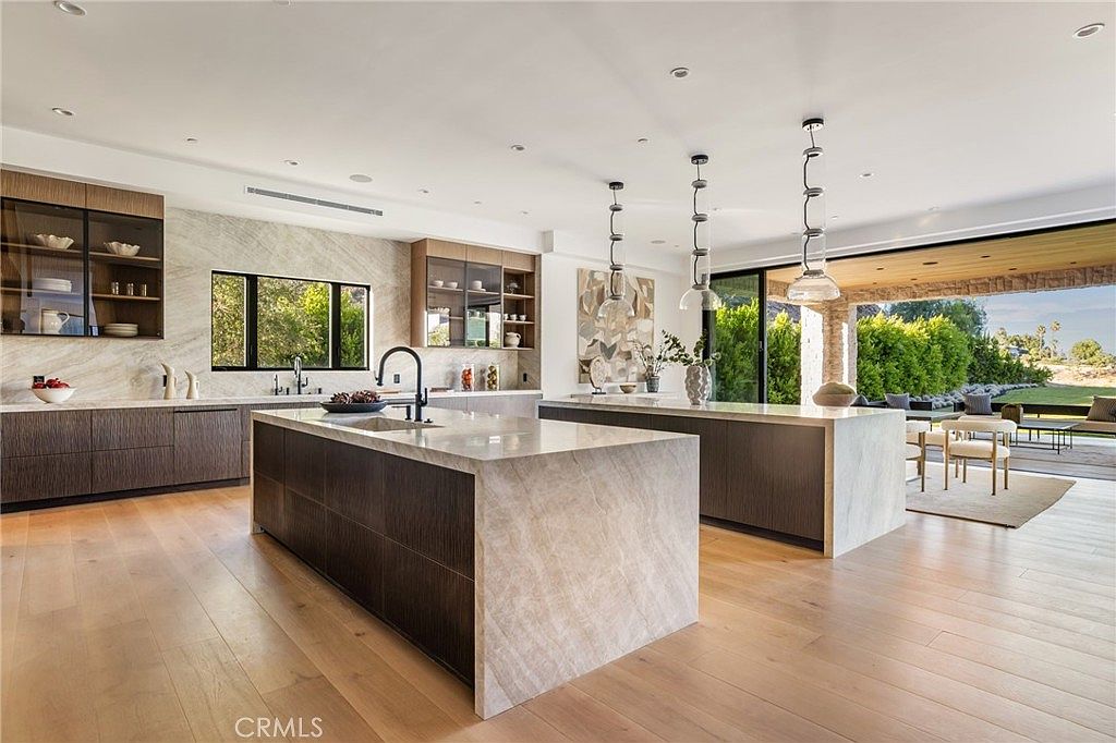 This is a bright and modern kitchen featuring two large islands with marble countertops and dark wood cabinetry. The kitchen is well-lit with pendant lighting and recessed lighting, and it opens to an outdoor living space through a large sliding glass door. The overall impression is one of luxury and spaciousness, perfect for entertaining.