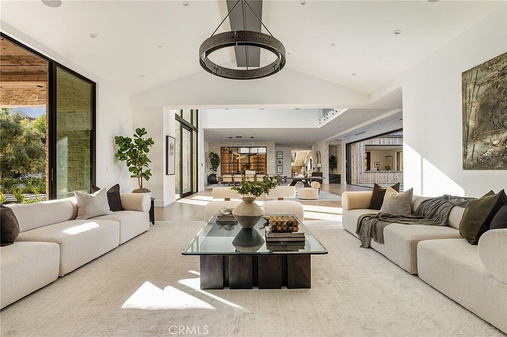 This is a spacious and bright living room featuring two large, light-colored sofas facing each other across a glass-topped coffee table. The room has high ceilings with a modern circular chandelier and large windows that let in ample natural light. The open floor plan leads to other areas of the house, creating a sense of flow and connectivity.