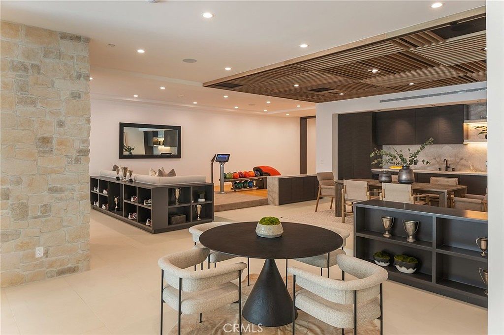 This interior shot showcases a modern living room with a unique blend of textures and design elements. A stone accent wall complements the sleek, dark cabinetry and shelving units, while a bowling alley setup adds a playful touch. The room features a round dining table with modern chairs, creating a stylish and inviting space.