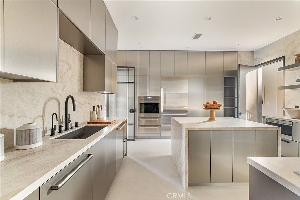 This is a modern kitchen with sleek, stainless steel cabinetry and a large island with a light-colored countertop. The kitchen features high-end appliances, including a built-in oven and refrigerator. The design is minimalist and sophisticated, with a focus on clean lines and neutral tones.