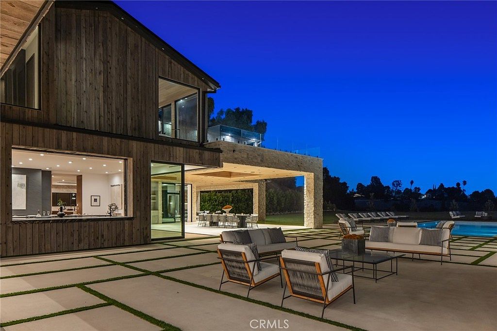 This image showcases a luxurious outdoor patio area at dusk, featuring modern furniture arrangements, including sofas and chairs, set on a paved patio with grass inlays. The patio extends from a contemporary home with large windows and a stone-covered overhang, leading to a dining area and a pool in the background. The scene exudes a sense of upscale outdoor living and relaxation.