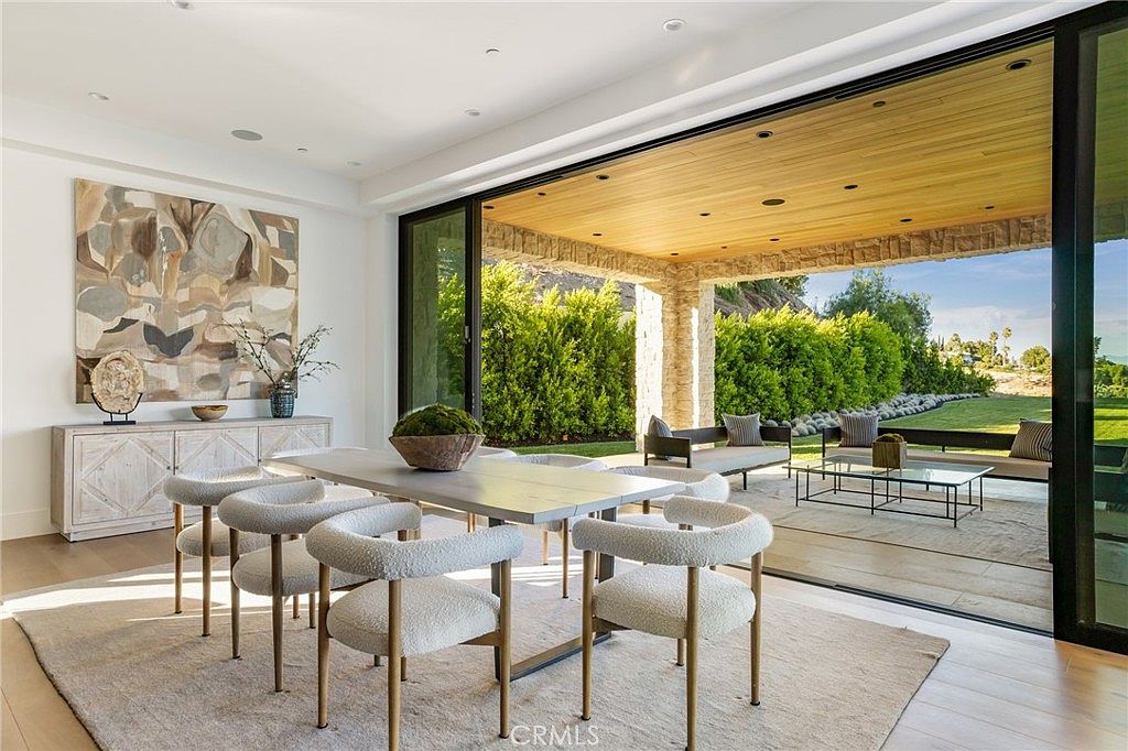 This is a bright and airy dining room featuring a modern aesthetic. The room includes a large dining table with six chairs, a sideboard with artwork above, and a large sliding glass door that opens to an outdoor patio area. The neutral color palette and natural light create a welcoming and sophisticated atmosphere.