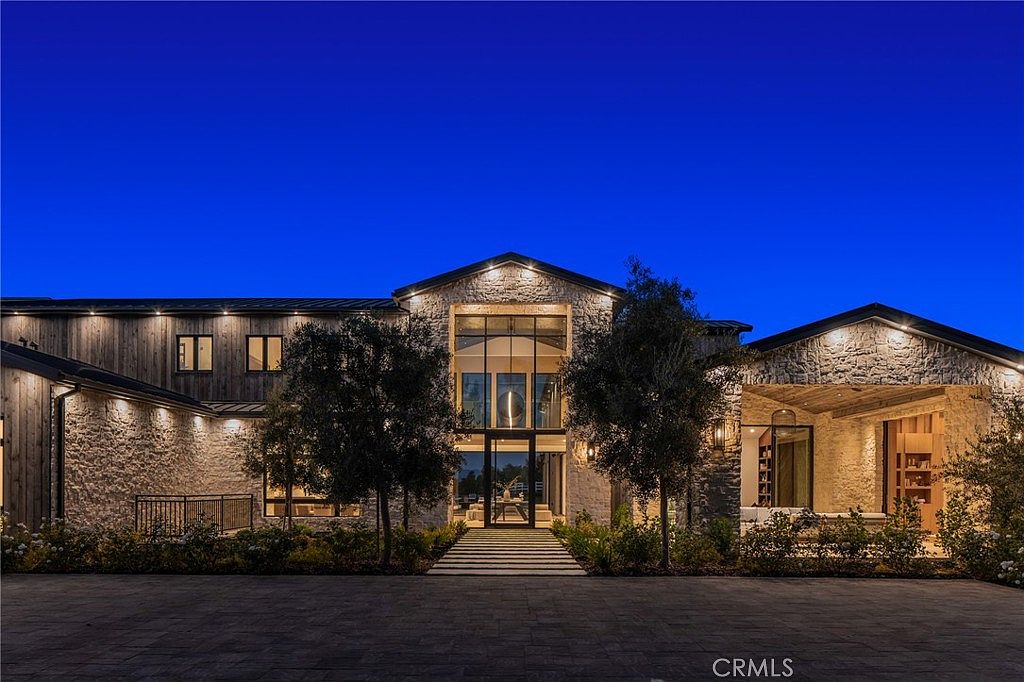 This is a front exterior view of a luxurious, modern farmhouse-style home at dusk. The facade features a combination of stone and wood siding, with a grand entrance highlighted by a large glass window and a series of steps leading up to the front door. The property is well-lit, showcasing the architectural details and landscaping.