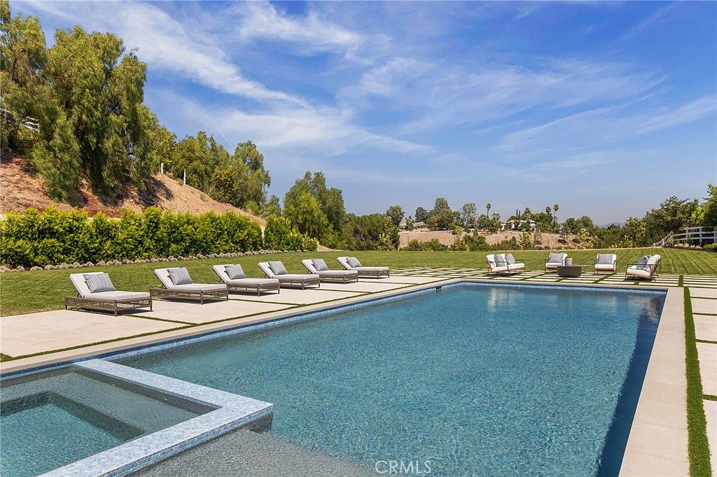 This image showcases a luxurious backyard pool and spa area. The pool features a clean, modern design with surrounding lounge chairs and a seating area with a fire pit. The landscape includes well-maintained grass and mature trees, creating a private and serene outdoor living space.