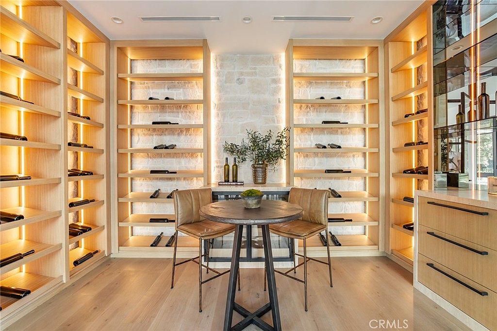 This is an interior shot of a well-appointed wine cellar. The room features custom wooden shelving filled with wine bottles, a stone accent wall, and a small round table with two chairs, creating an intimate tasting area. The lighting is warm and inviting, highlighting the rich textures and creating a luxurious ambiance.