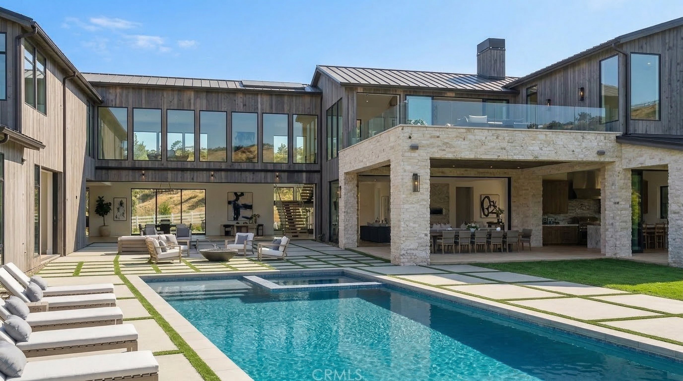 This stunning contemporary estate features a spacious rear courtyard highlighted by a large swimming pool and integrated spa. The home showcases a modern farmhouse aesthetic with striking vertical wood siding and a substantial stone-clad outdoor dining pavilion. The seamless integration between the indoor living spaces and the expansive patio area creates an ideal environment for luxury outdoor entertaining.