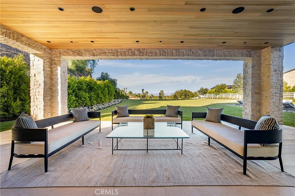This is a rear exterior view of a covered patio area, showcasing an outdoor living space. The patio features three sofas arranged around a glass coffee table, set on a large area rug. The structure is supported by stone pillars and has a wooden ceiling with recessed lighting, offering a seamless transition to the expansive lawn and landscape beyond.
