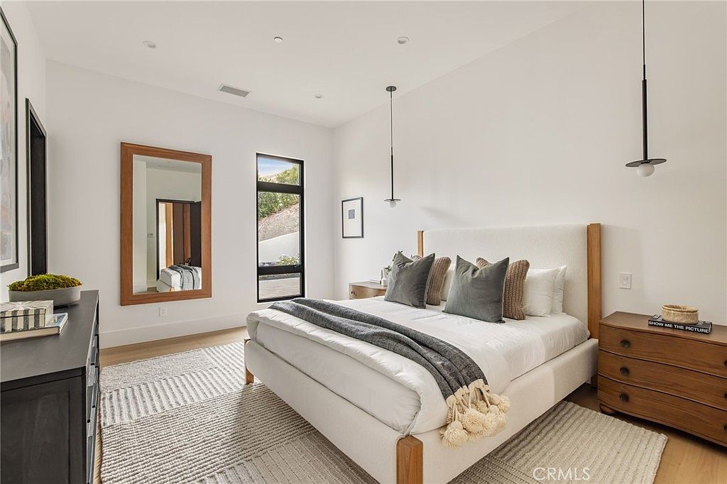 This is a bright and airy primary bedroom featuring a large bed with a white upholstered headboard and neutral-toned bedding. The room has a minimalist design with white walls, wood accents, and modern pendant lighting. A large mirror and a window add depth and natural light to the space, creating a serene and inviting atmosphere.