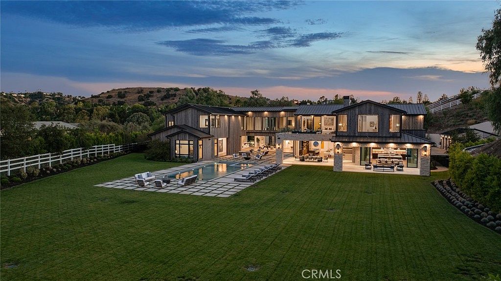 This is a rear exterior view of a large, modern home at dusk. The house features a pool with lounge chairs, a spacious lawn, and an expansive patio area with outdoor seating. The architecture is contemporary with a mix of wood and stone elements, creating a luxurious and inviting outdoor living space.