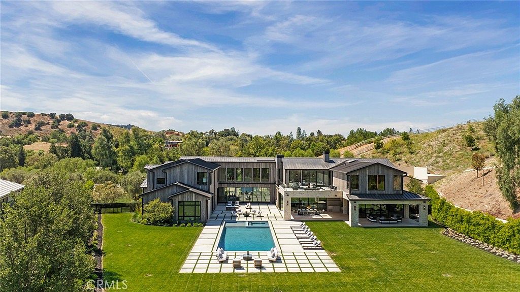 This is a rear view of a modern luxury home, showcasing a large swimming pool surrounded by a concrete patio with lounge chairs and outdoor seating. The house features a contemporary design with large windows and a covered patio area, all set against a backdrop of lush greenery and rolling hills. The manicured lawn and landscaping enhance the property's appeal.
