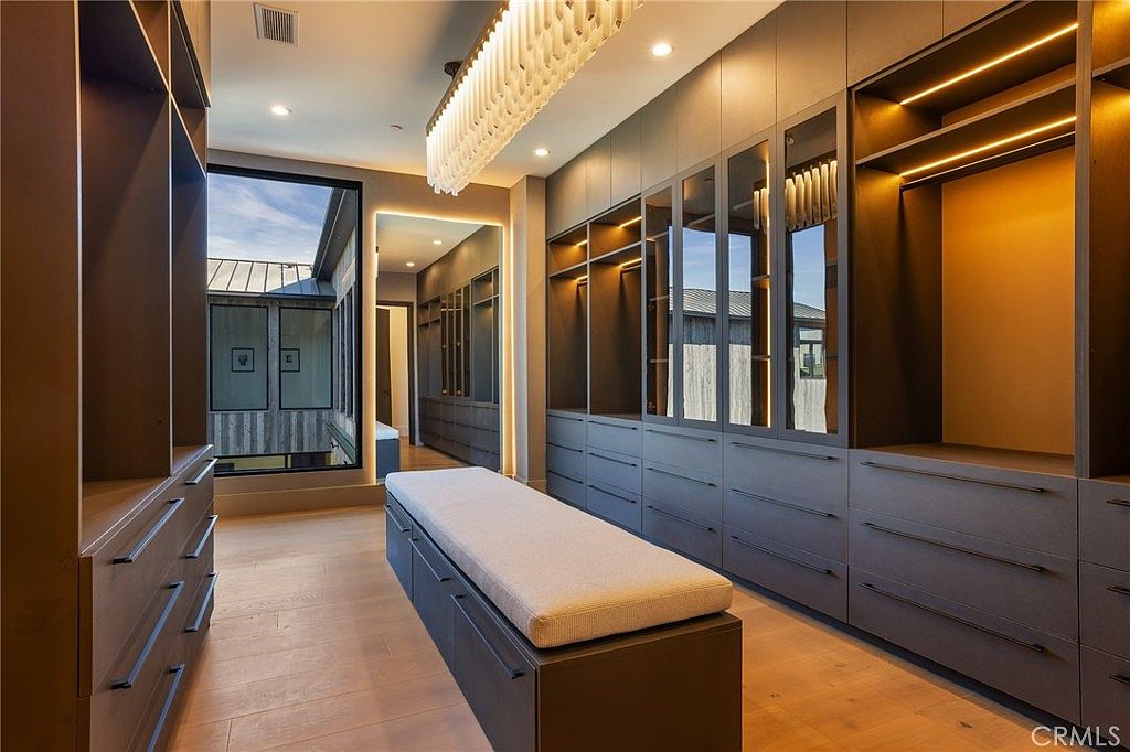 This is a well-organized walk-in closet featuring custom cabinetry with drawers and shelving, some with glass fronts. A central bench with a cushioned top provides a seating area. The closet is illuminated by a modern chandelier and recessed lighting, creating a luxurious and functional space.