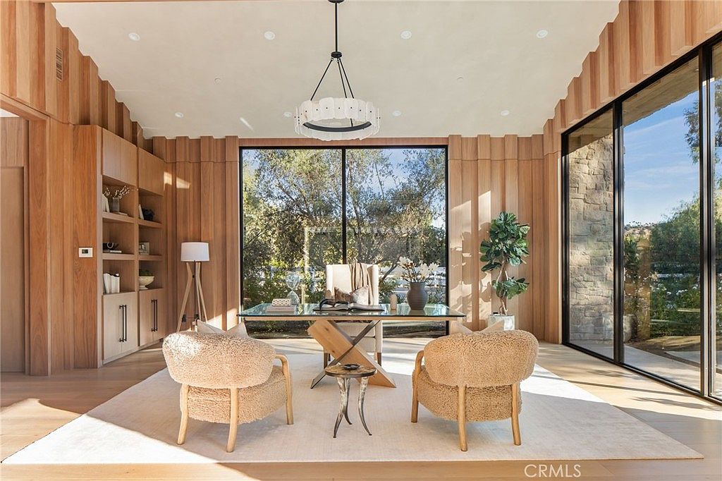 This is an interior shot of a modern office space featuring wood-paneled walls and large windows offering a view of the outdoors. The room is furnished with a glass-top desk, two plush chairs, and a unique side table, all set on a light-colored rug. A contemporary chandelier hangs from the ceiling, adding to the room's sophisticated and inviting atmosphere.