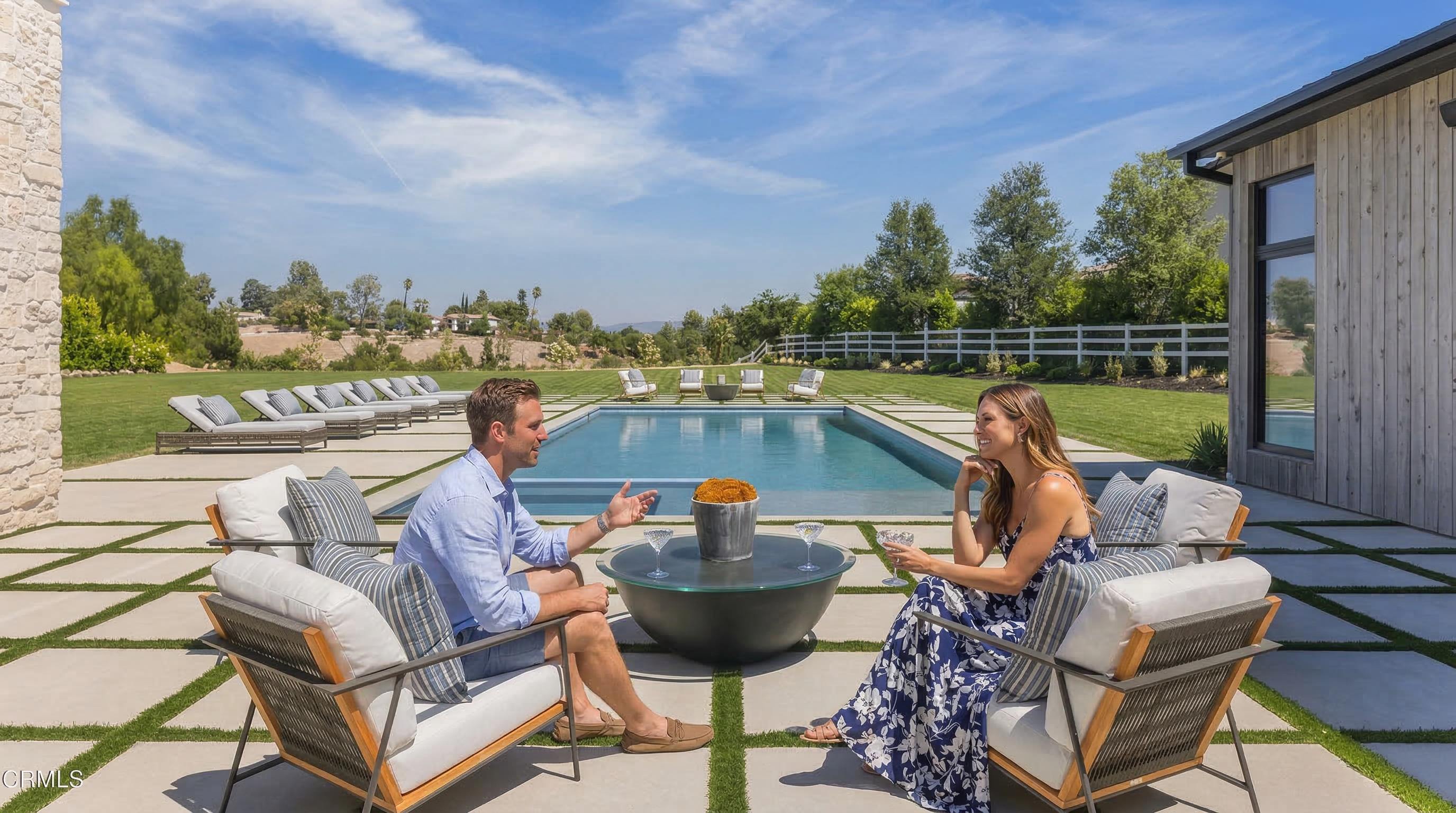 This stunning outdoor living space features a contemporary stone-tiled patio surrounding a rectangular swimming pool, perfect for entertaining. The area is furnished with a modern circular fire pit table and chic lounge chairs, complemented by a serene backdrop of manicured lawns and a white fence. The overall aesthetic is one of elegant, sun-drenched relaxation and high-end residential luxury.