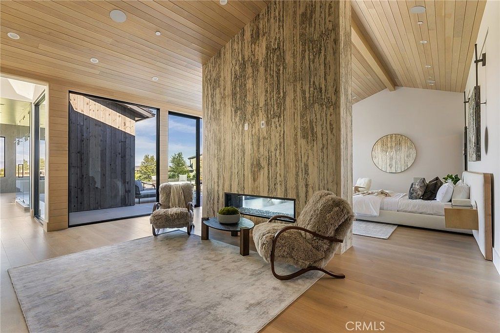 This is a luxurious primary bedroom featuring a modern fireplace and a serene sitting area with two plush chairs. The room boasts light wood flooring and ceiling, complemented by a neutral color palette. A large sliding glass door provides access to the outside, enhancing the room's spacious and airy feel, while a round mirror adds a touch of elegance above the bed.