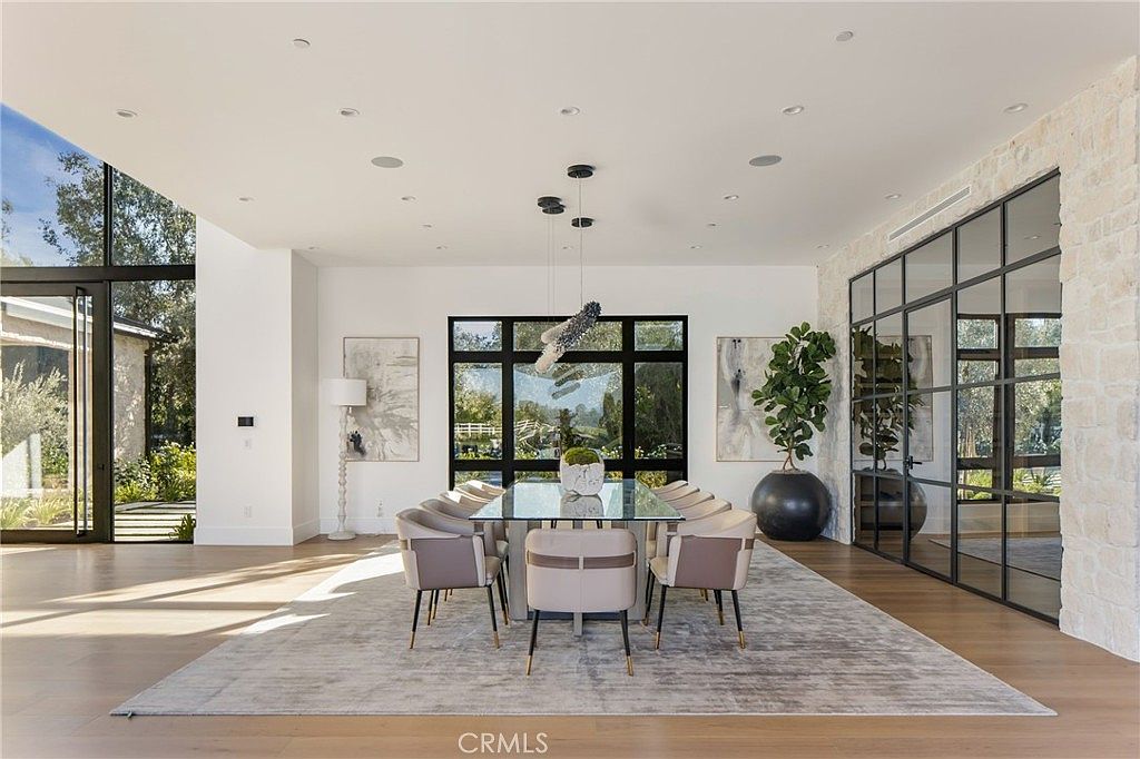 This is a bright and spacious dining room featuring a large glass-top dining table surrounded by modern upholstered chairs with gold accents. The room is well-lit with natural light from large windows and a contemporary chandelier. A neutral-toned rug anchors the space, and artwork and a large potted plant add to the room's sophisticated ambiance.