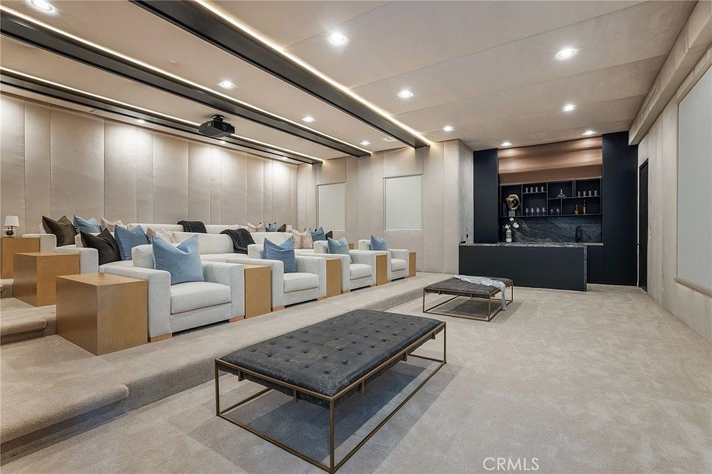This is a luxurious home theater featuring tiered seating with plush armchairs, each adorned with decorative pillows. A sleek bar area with dark cabinetry and marble countertops is located at the back of the room, adding a touch of sophistication. The room is well-lit with recessed lighting and accent lights, creating a comfortable and inviting atmosphere for entertainment.