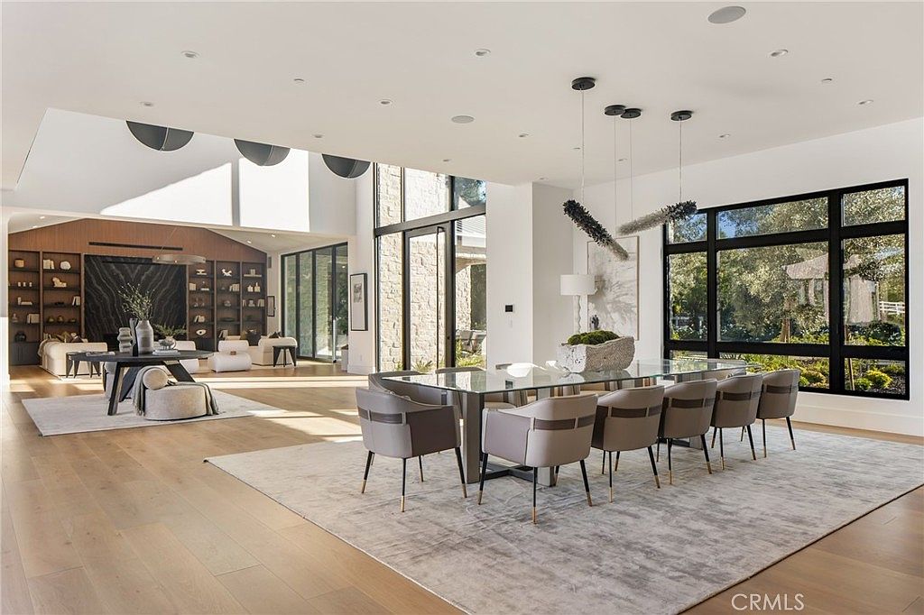 This is a bright and spacious dining room featuring a large glass-top dining table surrounded by modern chairs with sleek black legs. The room is well-lit with natural light from large windows and stylish pendant lighting above the table. A neutral-toned rug anchors the dining area, and the open floor plan connects to a living space with built-in shelving, creating a seamless flow.