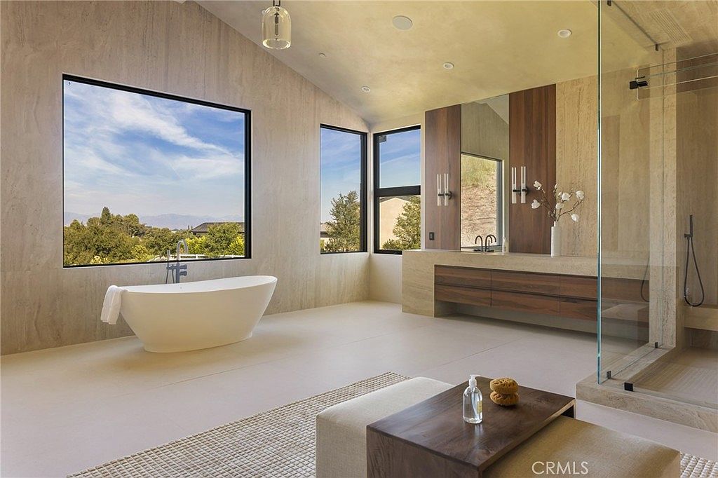 This is a luxurious primary bathroom featuring a freestanding bathtub positioned beneath a large window offering scenic views. The space includes a modern vanity with wood accents, a glass-enclosed shower, and neutral-toned tile flooring, creating a spa-like atmosphere. The perspective is from a low angle, emphasizing the spaciousness and high ceilings.