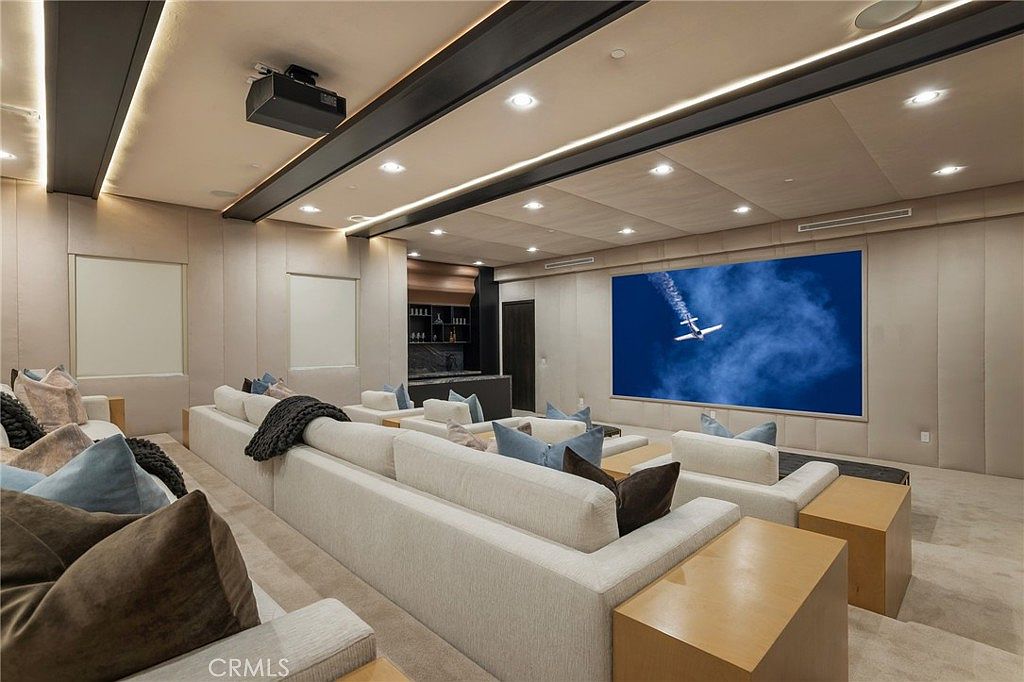 This is an interior shot of a home theater, featuring tiered seating with plush, light-colored sofas and throw pillows. A large screen displays an image of an airplane, and a projector is mounted on the ceiling. The room also includes a bar area in the back, creating a luxurious and immersive entertainment space.