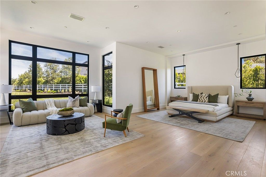 This is a spacious primary bedroom featuring a seating area with a modern sofa and a unique coffee table, complemented by a large window offering natural light. The sleeping area includes a platform bed with stylish pillows and a bench, flanked by nightstands and pendant lighting. The room is decorated in neutral tones with hardwood flooring and area rugs, creating a serene and luxurious atmosphere.